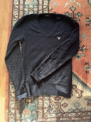 Mörkblå kabelstickad tröja GANT - Mörkblå kabelstickad tröja från GANT med V-ringning och broderad logga på bröstet. Tröjan har långa ärmar, ribbade muddar och klassisk passform. Perfekt för en stilren och tidlös look.