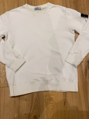 Vit långärmad tröja från Stone Island - Vit långärmad sweatshirt från Stone Island med klassisk rund hals och ribbade muddar. Tröjan har en enkel design och en svart logotyp-patch på vänster ärm. Perfekt för en clean och stilren look.