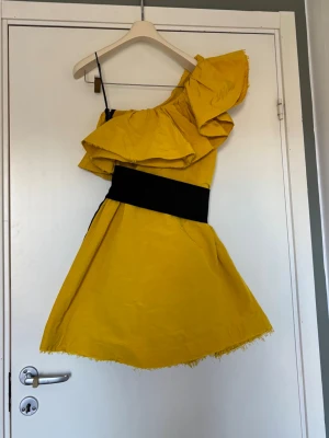 Gul oneshoulder klänning med volang - Snygg gul kortklänning med stor volang upptill och svart midjeband. Klänningen har oneshoulder-design. Jag har själv köpt den här på Plick och den har tyvärr inget märke i sig 