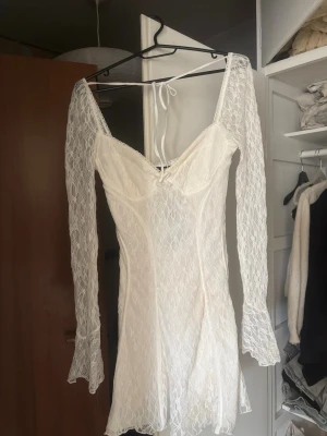Vit spetsklänning med v-ringning - Säljer en vit kort spetsklänning med v-ringning och långa, lätt utsvängda ärmar. Klänningen har snörning i ryggen och är figurnära med fina spetsdetaljer över hela plagget. Perfekt för studenten, midsommar eller sommarfester. Nyköpt från New Yorker knappt använd, prislappen sitter fortfarande kvar. 