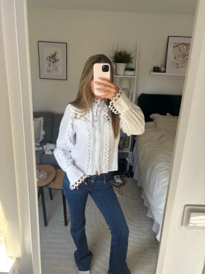 Vit blus med spetsdetaljer - Superfin vit blus med långa ärmar och dekorativa spetsdetaljer längs knäppningen, axlarna och ärmsluten. Blusen har en klassisk krage och är tillverkad i ett lätt tyg som ger en romantisk och trendig look.