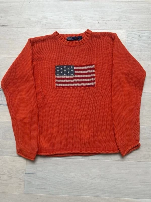 Polo Ralph Lauren Flag Sweater – Ikonisk vinterklassiker i statementfärg  - Polo Ralph Lauren Flag Sweater – Ikonisk vinterklassiker i statementfärg 🔥  Nu har du chansen att lägga vantarna på en av Polo Ralph Laurens mest klassiska modeller – den stickade tröjan med den ikoniska amerikanska flaggan framtill. En tidlös design som andas preppy, kvalitet och exklusivitet.  Den klara orange färgen gör att du verkligen sticker ut i vintermörkret, samtidigt som den grovstickade bomullen ger en varm och gedigen känsla – perfekt för kalla dagar. Matcha med jeans för en avslappn