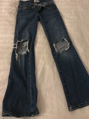 Blå slitna jeans från 157 - Säljer ett par blå jeans från 157 med stora slitningar över knäna och klassisk femficksmodell. Jeansen är bootcut och är gjorda i denim med en snygg tvätt. dessa har bara använts ca 10 gånger. 
