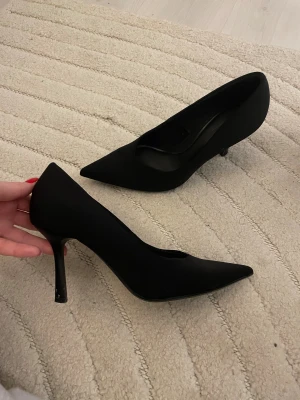 Svarta spetsiga pumps med smal klack - Säljer ett par klassiska svarta pumps med spetsig tå och smal hög klack. Skorna har en elegant siluett och är tillverkade i ett matt material som ger en stilren look. Perfekta för dig som vill ha en tidlös och trendig sko till garderoben. 
