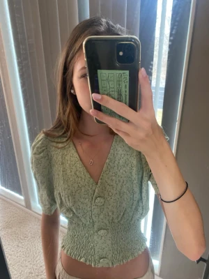 Grön blommig blus med knappar - Söt ljusgrön blus med små blommor, croppad passform och smockad midja. V-ringad med stora knappar framtill och korta puffärmar. Perfekt för vår och sommar, supergullig och snygg. 