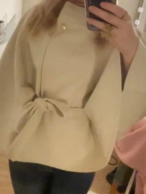 beige kappa från nelly - Snygg beige cape-kappa med knytband i midjan och en dekorativ knapp vid halsen. Kappan har vida ärmar och en stilren, minimalistisk design som passar perfekt till jeans eller dressade byxor. Perfekt för dig som vill ha en trendig och unik look.