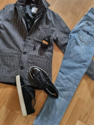 Grå overshirt från Bläck - Stilren grå overshirt från Bläck i mjukt tyg med diskret fiskbensmönster. Klassisk krage, tre framfickor och knäppning med svarta knappar. Perfekt lager-på-lager-plagg med modern passform och snygga detaljer.