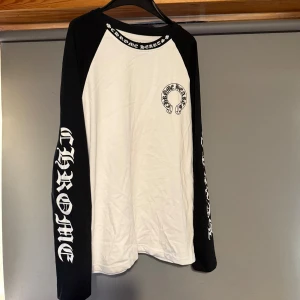 Chrome Hearts långärmad tröja med tryck - Säljer en långärmad svartvit tröja från Chrome Hearts med stora tryck på ryggen och ärmarna. Tröjan har svart ärmar och vit kropp, samt logga på bröstet och text längs båda ärmarna. Perfekt för dig som gillar streetwear och tydliga loggor.