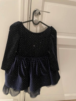 Mörkblå sammetsklänning med rosett - Supersöt mörkblå sammetsklänning med vita prickar och stor rosett bak. Klänningen har lång ärm, puffiga axlar och volangkant nertill. Perfekt för dig som vill sticka ut lite extra! Strl 128