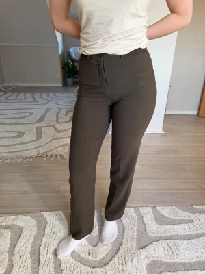 Bruna kostymbyxor från  - Snygga bruna kostymbyxor från Mari Philippe med raka ben och hög midja. Byxorna har klassisk dragkedja och knapp framtill samt bälteshällor. 