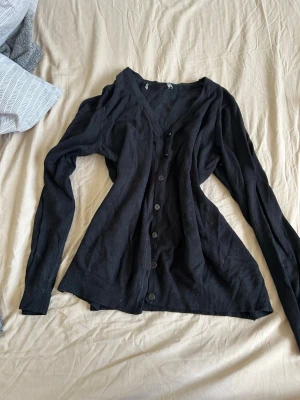 Svart tunn kofta med knappar - En svart, tunn och långärmad kofta med v-ringning och knäppning framtill. Koftan har ribbade muddar vid ärmslut och nederkant, perfekt att slänga på sig över en t-shirt eller linne. Enkel och stilren design som passar till det mesta.