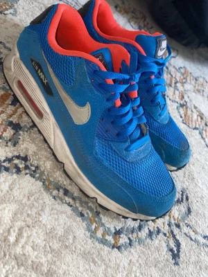 Nike Air Max blå sneakers - Nike Air Max sneakers i blått mesh och mocka med vita och grå detaljer samt röd insida. Klassisk Air Max-sula med synlig luftbubbla och tydlig Nike-logga på sidan. Skorna har blå skosnören och en sportig vibe som sticker ut.