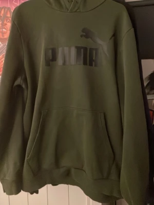 Olivgrön hoodie från Puma - Säljer en olivgrön hoodie från Puma med stor svart logga på bröstet och klassisk känguruficka. Hoodien har huva och ribbade muddar vid ärmslut och nederkant. Perfekt för dig som gillar streetwear och sportig stil.