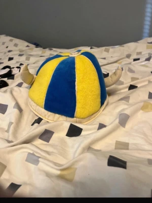 Vikingahjälm i blått och gult - Säljer en mjuk vikingahjälm i plysch med blå och gula paneler samt två vita horn på sidorna. Perfekt accessoar för att sticka ut på matchen eller temafesten. Hjälmen har en rund form och är riktigt bekväm att bära.