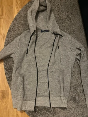 Äkta Polo Ralph Lauren - Snygg grå hoodie med dragkedja från Polo Ralph Lauren. Klassisk design med svart logga broderad på bröstet, huva med snören och två fickor framtill. Perfekt för en avslappnad och stilren look.Äkta Ny pris 1200
