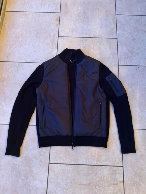 Svart cardiganjacka från J.Lindeberg - Stilren svart cardiganjacka från J.Lindeberg med stickade ärmar och quiltad front. Jackan har dragkedja framtill, ribbade muddar och en liten logga på bröstet. Perfekt för lager-på-lager och snygg till jeans.