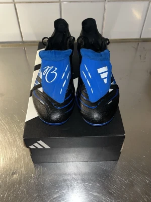 Adidas Predator fotbollsskor blå/svart - Snygga Adidas Predator fotbollsskor i svart och blått med vita detaljer. Skorna är även gjorda för Jude Bellingham. Skorna har en modern design med täckt snörning och tydliga Predator-loggor på sidan. Ovandelen är i syntetmaterial och sulan har fasta dobbar för spel på gräs. Pris kan diskuteras och även byten med andra skor kan funka.