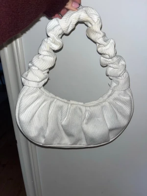 Vit scrunchie-handväska i tyg - Trendig handväska i vit ribbad textil med scrunchie-inspirerat handtag. Väskan har en mjuk, rundad form och är perfekt för att lyfta din outfit. Ingen synlig logga eller märke. Materialet känns mjukt och väskan har en modern vibe.