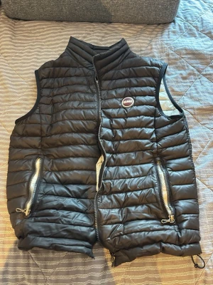 Svart dunväst från Colmar - Snygg svart dunväst från Colmar med quiltad design och två dragkedjeförsedda fickor framtill. Västen har hög krage och Colmar-logga på bröstet. Insidan är ljusbeige och västen har justerbar nederkant med dragsko. Perfekt för lager-på-lager och streetstyle.