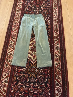 Grå velourbyxor Juicy Couture - Säljer ett par grå mjukisbyxor från Juicy Couture i mjuk velour. Byxorna har raka ben, bred resår i midjan med dragsko och stora fickor framtill. Klassisk Juicy Couture-logga broderad på ena fickan. Perfekta för en chill och trendig look.