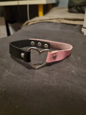 Svart och rosa choker med hjärta - Snyggt choker i konstläder med ena sidan svart och andra sidan rosa. Chokern har ett metallhjärta i mitten och tryckknappar för justering. Perfekt accessoar för dig som gillar alternativ stil och vill sticka ut lite extra.
