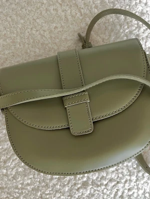 Axelremsväska i ”matchalattegrön” - En axelremsväska i en dov ”matchalattegrön nyans”. Väskan har en minimalistisk design som passar till det mesta, men den har spår av användning som jag vill vara tydlig med.  Väskan är i ett fint helhetsläder-liknande material, men det finns synliga skavmärken/streck på locket (se närbilderna). Dessa syns i vissa vinklar men påverkar inte väskans funktion.  Det finns ett mindre fack med dragkedja inuti för värdesaker. Justerbar axelrem och dekorativt spänne framtill. Väskan stängs med magnetlås 