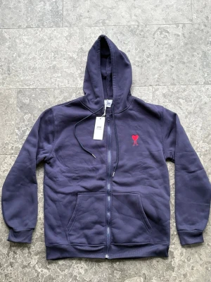 Mörkblå hoodie med dragkedja från Ami - Snygg mörkblå hoodie från Ami med dragkedja och huva. På bröstet finns Ami:s ikoniska röda hjärta med bokstaven A broderat. Hoodien har känguruficka och ribbade muddar. Tillverkad i mjuk bomull för maximal komfort.