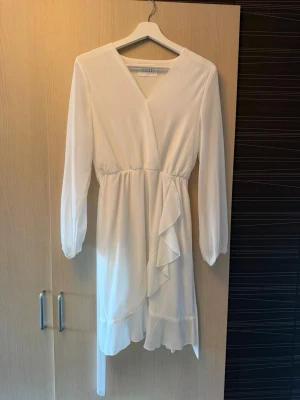 Vit omlottklänning med volang - Snygg vit omlottklänning från NA-KD med långa ärmar och volangdetalj framtill. Klänningen har v-ringning och markerad midja med knytband. Perfekt för sommarens alla tillfällen. Endast använd en gång! 💘