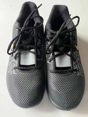 Nike Kyrie svart/guld sneakers - Snygga Nike Kyrie sneakers i svart mesh med guldiga detaljer och logga på hälen. Skorna har lågt skaft, svarta snören och mönstrad ovandel för extra stil. Perfekta för dig som gillar basketinspirerad design och vill sticka ut med coola detaljer.