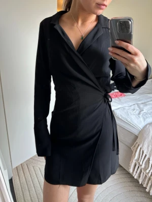 Svart omlottklänning NA-KD - Snygg svart omlottklänning från NA-KD med långa ärmar och knytband i midjan. Klänningen har en kort längd och klassisk krage, perfekt för en trendig och stilren look.