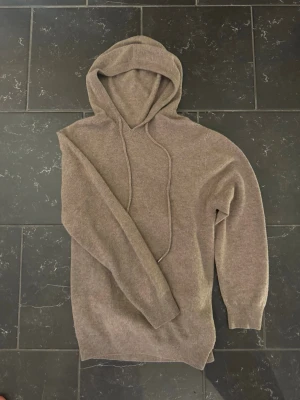 Beige stickad hoodie med huva - Mysig beige stickad hoodie med huva och dragsko. Tröjan har ribbade muddar vid ärmslut och nederkant, och är tillverkad i ett mjukt material som passar perfekt för en avslappnad stil. Enkel och stilren design utan synliga loggor eller tryck.