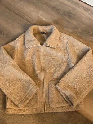 Beige teddyjacka med dragkedja - Mysig beige teddyjacka i fluffigt material med klassisk krage och hel dragkedja framtill. Jackan har två stora fickor och raka ärmar. Perfekt för dig som gillar en avslappnad och trendig look. Materialet är mjukt och värmande, perfekt för kyliga dagar.