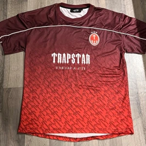 Trapstar röd t-shirt - Säljer denna snygga limited editon t-shirt från Trapstar i röd och vinröd ton med vit logga och text framtill. Tröjan har ett sportigt tryck med siffran 22 på ryggen. Storlek S men passar också M