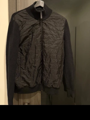 Svart Cardigan från J.Lindeberg - Snygg svart cardigan från J.Lindeberg med quiltad front och stickade ärmar samt rygg. Jackan har hög krage, dragkedja framtill och två sidofickor med dragkedja. Perfekt för kalla dagar och ger en modern look.