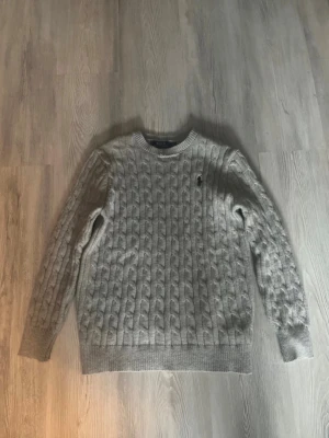 Grå kabelstickad tröja från Polo Ralph Lauren - Snygg grå kabelstickad tröja från Polo Ralph Lauren med klassisk rund halsringning och svart broderad logga på bröstet. Storlek S,