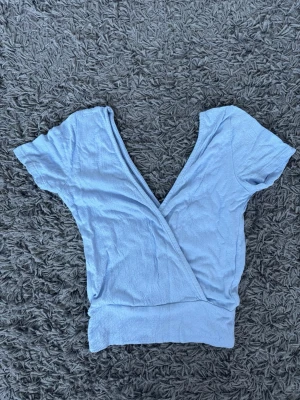 Ljusblå omlottopp från Stradivarius storlek XS - Supersöt ljusblå omlottopp från Stradivarius med korta ärmar och ribbad struktur. Toppen har en djup v-ringning och bred linning nedtill som ger en snygg passform. Perfekt att styla med jeans eller kjol för en trendig look.