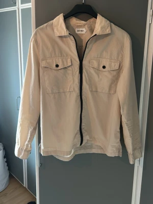 Beige overshirt från East West - Stilren beige overshirt från East West med två bröstfickor med lock och knapp, dragkedja framtill och klassisk krage. Tillverkad i ett lätt bomullstyg som passar perfekt för lager på lager. Långärmad med knappar vid ärmslut för extra detalj.