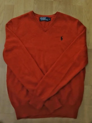 Röd V-ringad tröja från Polo Ralph Lauren - Säljer en röd v-ringad tröja från Polo Ralph Lauren med klassisk broderad logga på bröstet. Tröjan har lång ärm och är tillverkad i mjuk ull som känns skön mot huden. Perfekt för dig som gillar stilrena och tidlösa plagg. Axelbredd: 43cm / Längd: 63cm. Nypris: 725kr / Mitt pris: 189kr