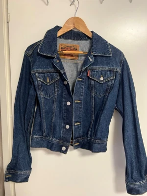 Mörkblå jeansjacka från Crocker - Klassisk mörkblå jeansjacka från Crocker med silverfärgade knappar och två bröstfickor med lock. Jackan har en rak passform, markerad krage och detaljerade sömmar. Perfekt för en avslappnad och cool stil. Ikonisk röd Crocker-tagg på bröstfickan.