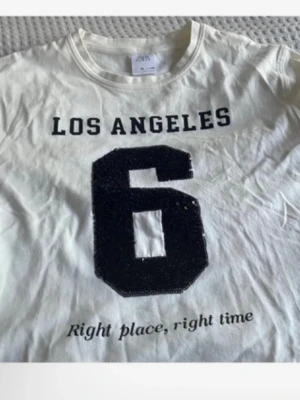 Vit t-shirt med tryck från Zara - Vit t-shirt från Zara med svart text 'LOS ANGELES', stor siffra 6 och texten 'Right place, right time' framtill. Klassisk rund hals och korta ärmar. Tillverkad i mjuk bomull, perfekt för en avslappnad streetstyle-look.