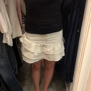 Vit volangkjol från Gina tricot - Supersöt vit kortkjol från Gina med två lager volanger och råa kanter för en cool, avslappnad vibe. Klassisk jeansmodell med fickor fram samt bälteshällor. Perfekt för dig som gillar en trendig och lekfull stil.