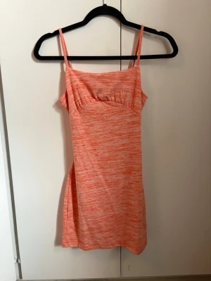 Kort orange klänning med knyt - Snygg kort klänning i orange och vitt melerat tyg med smala axelband och knyt i ryggen. Klänningen har en figurnära passform och är perfekt för varma dagar. Materialet känns mjukt och stretchigt. Endast testad.