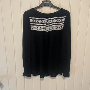Svart blus med broderi från Paramount - Svart blus från Paramount med vita broderade detaljer över bröstet. Blusen har rund halsringning och långa ärmar. Materialet känns mjukt och luftigt, perfekt för en avslappnad men ändå stilren look.