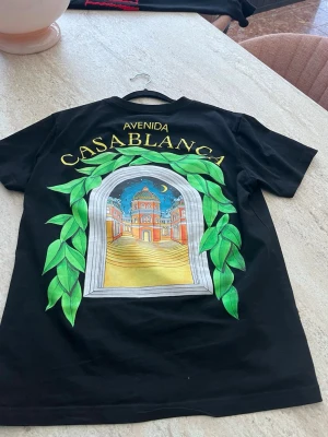 Svart Casablanca Avenida t-shirt - Säljer en svart t-shirt från Casablanca med stor färgglad grafik på ryggen. Trycket visar en fönsterliknande ram med gröna blad och en byggnad i solnedgång, samt texten 'Avenida Casablanca' i gult. T-shirten har rund hals och korta ärmar.