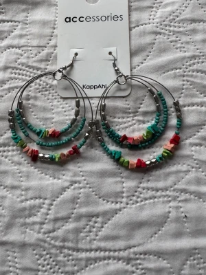 Nya. Färgglada creolörhängen KappAhl - Stora creolörhängen från KappAhl med pärlor i turkos, silver, rött, grönt och rosa. Örhängena har flera rader med små pärlor och oregelbundna stenar för en bohemisk vibe. Perfekta för dig som gillar färg och unika detaljer.