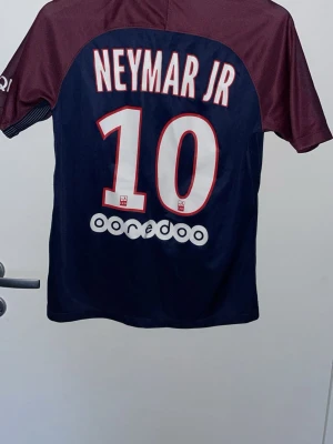 PSG Neymar JR matchtröja Nike - Säljer en mörkblå och vinröd PSG matchtröja med Neymar JR och nummer 10 på ryggen. Tröjan har korta ärmar, Fly Emirates-tryck på bröstet och Nike-logga. Barn storlek XL