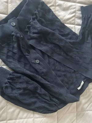 Svart kofta från Jacqueline de Yong - Mysig svart kofta från Jacqueline de Yong med snyggt strukturmönster och stora ribbade muddar. Koftan har knäppning framtill med 4 knappar och ballongärmar