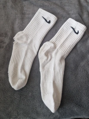 Vita ribbade Nike strumpor - Säljer ett par klassiska vita ribbade strumpor från Nike med svart broderad logga upptill. Strumporna är i bomullsblandning och har en sportig look med förstärkt tå och häl. Perfekta till sneakers eller träning. Hål i de men går o sy