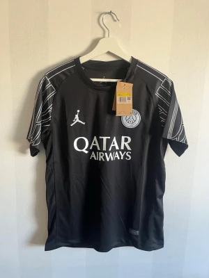 Svart PSG x Jordan matchtröja - Snygg svart PSG matchtröja i samarbete med Jordan. Tröjan har vita detaljer och tryck, inklusive Qatar Airways-logga och PSG-emblem på bröstet. Ärmarna har coola vita linjemönster. Perfekt för fotboll eller träning.