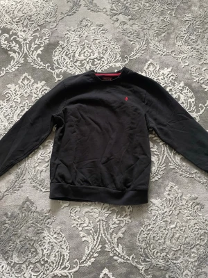 Svart sweatshirt från MCN - Klassisk svart sweatshirt från MCN med rund halsringning och diskret röd logobrodyr på bröstet. Tröjan har långa ärmar och ribbade muddar vid ärmslut och nederkant. Perfekt till jeans eller joggers för en avslappnad look.